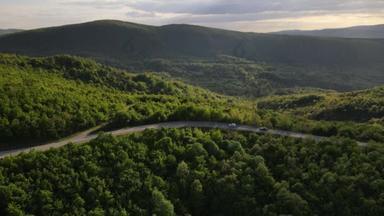 在塞尔维亚，春车沿着蜿蜒的<strong>公路</strong>行驶时，在<strong>山脉</strong>上一条弯曲的沥青路面的空中无人驾驶镜头，周围环绕着绿地和景观，交通和旅行的概念