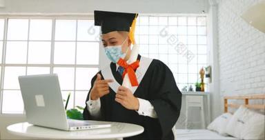 头戴医疗面具，头戴笔记本电脑，坐在办公桌前，拿着毕业文凭，喜庆成功的大学生或大学生