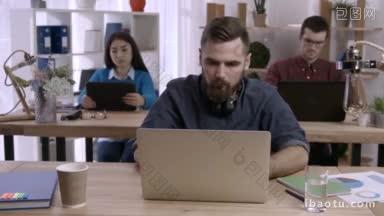 压力潮人胡子工作在<strong>桌子上</strong>的笔记本电脑在繁忙的创意办公室和皱褶纸筋疲力尽的商人下