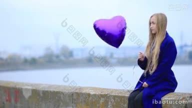 美丽悲伤的金发女人坐在胸墙上，拿着紫色的心<strong>气球</strong>在城市的背景独自在情人节