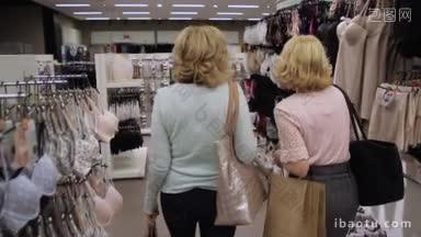 后视图的两个迷人的金发女人拿着购物袋走过货架上的时装服装店不同的衣服