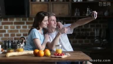 迷人的<strong>年轻情侣</strong>坐在厨房餐桌边吃早餐，一边用智能手机自拍，一边微笑