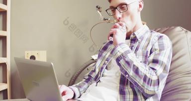 呼气烟电子烟工作移动PC年轻的男人。放松工作过程坐着沙发工作首页距离自由职业者男人。首页办公室
