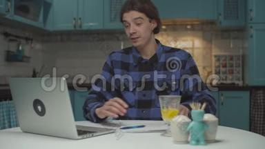 年轻的20多岁的商人通过笔记本电脑在线交谈，并在蓝色厨房上展示纸质文件。 有纸的男企业家