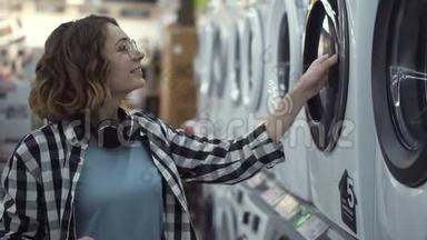 一个年轻的积极的女人穿着格子衬衫，在<strong>家用电器</strong>商店里选择洗衣机。 打开门然后