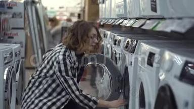 一个年轻的积极的女人穿着格子衬衫，在家用电器商店里选择洗衣机。 打开门然后