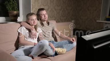 年轻可爱的夫妇坐在客厅里拥抱，看电视，吃爆米花。