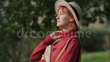 年轻迷人的红头发女人，戴着帽子和毛衣，在自然公园的长凳上看书。 秋天凉爽的一天。