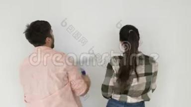 这对年轻夫妇画了一堵白色的墙。 男人和女孩站在一个房间的后面，手里拿着滚轮