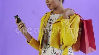 非洲裔美国妇女的肖像与购物袋和手机紫色背景在工作室。 黄色夹克
