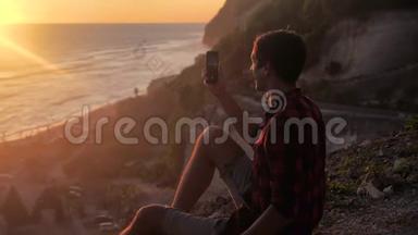 年轻人的剪影在他的智能手机上制作美丽日落的照片或视频。 旅游博主制作社交照片