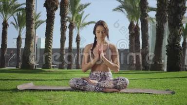 漂亮的女人坐在草地上的莲花姿势。 在公园练习瑜伽的教练