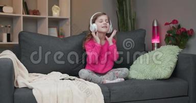 小女孩在沙发上跳舞，在家听耳机里的音乐。 戴着耳机跳舞唱歌唱歌的女孩