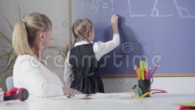 白人小女孩在黑板上写数学公式。 坐在餐桌前的中年教师