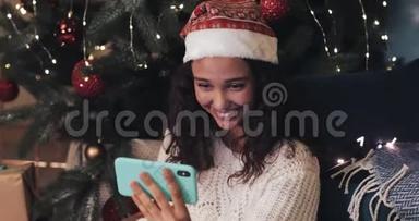 戴着圣诞帽的年轻穆拉托女孩微笑着坐在圣诞树下，用她的现代手机打视频电话