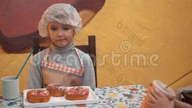 可爱的女孩戴着厨师帽，吃甜甜圈，从杯子里喝可可。 小女孩用吸管吃甜甜圈