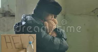 一个孤独的无家可归的孩子穿着脏衣服坐在帮助纸板和拥抱泰迪熊的肖像。 绝望