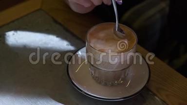 年轻女子小心翼翼地把咖啡和泡沫慢慢地混合在一个玻璃杯里，坐在桌边的咖啡馆里。
