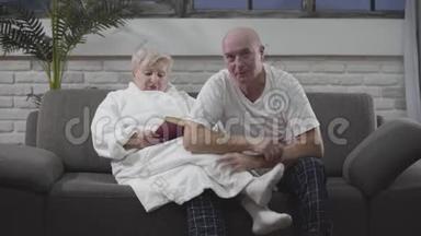 成熟的白人男人和妻子坐在沙发上看电视上的体育比赛。 女人在看书。 老年人休息