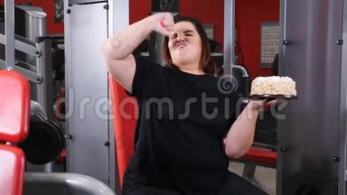 暴饮暴食的概念。女肥胖女子在健身房拿着<strong>大</strong>蛋糕，拇指朝下。<strong>搞</strong>笑胖女孩坐健身