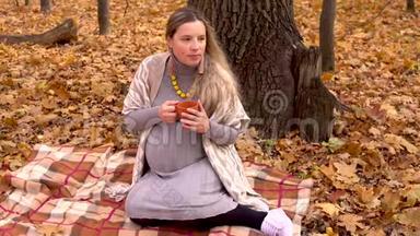一位孕妇坐在一棵树下，在秋天的森林里，坐在格子布上，喝着热茶。 黄色的叶子