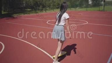 穿着运动鞋，裙子和t恤的少女在户外篮球场上骑着黄色滑板.. 影子跟随着