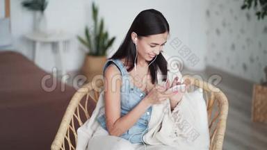 戴耳机的年轻女子坐在家里的椅子上，用电话上网聊天
