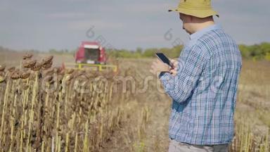 英俊的农民与智能手机站在田间向日葵与联合收割机的背景。 现代概念