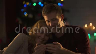 可爱的一对在闪闪发光的圣诞树下在智能手机上看电影，亲密