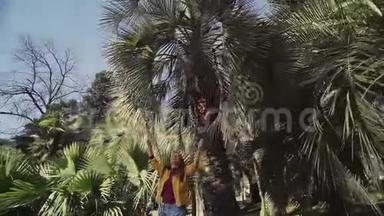 一个穿着黄色夹克衫的开朗的女孩正在玩棕榈叶，在阳光明媚的日子里在棕榈树<strong>下</strong>旋转。