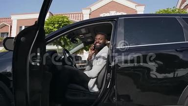 年轻的非洲商人西装革履，坐在<strong>新</strong>车的轮子上，在家附近停车，一边在智能手机上交谈。 多莉
