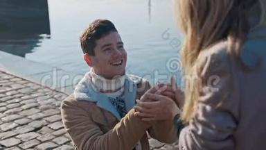英俊的恋爱中的年轻人单膝站立，拿着戒指，向码头上的女朋友求婚。 爱，家庭