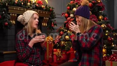 两个穿着红色格子衬衫的女孩坐在圣诞树<strong>下</strong>，戴着温暖的白色和紫色的帽子，认真地说话