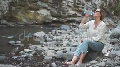 一位年轻漂亮的<strong>游</strong>客在靠近山河的石头上，喝着波约米<strong>温泉</strong>里清澈晶莹的清水