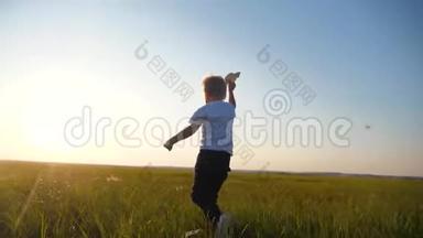 一个金发小男孩跑过绿色新鲜的草地，手里拿着一架纸飞机。 孩子梦想飞翔
