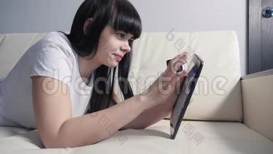 女孩用数字平板电脑制作网上销售购物概念。 黑发女人躺在沙发上看着网上商店，通过支付