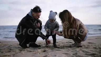 爸爸妈妈带着孩子蹒跚学步的小男孩在寒冷、多云、多风的天气里玩沙子，男人帮助小男孩