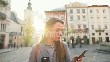 年轻的女人手里拿着一个保温杯，在日落时用智能手机沿着一条老街走着。 交流、社会