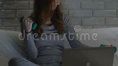 女人在床上用哑铃训练，在笔记本电脑上看健身视频，锻炼身体