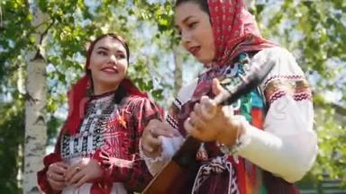 两个穿着俄罗斯服装的微笑女人站在桦树<strong>下</strong>，和巴拉莱卡一起唱歌