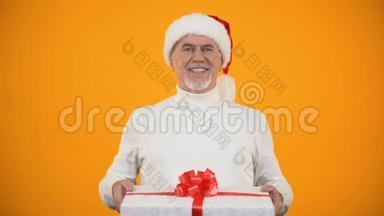 戴着圣诞老人礼帽的慈祥的老人在镜头前展示礼品盒和微笑的惊喜