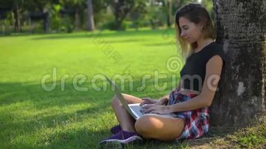 漂亮的成年女孩<strong>坐在</strong>公园的<strong>树下</strong>，把电脑放在膝盖上，