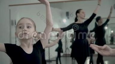 穿着黑色紧身衣的欧洲金发小芭蕾舞女与老师和其他女孩一起做芭蕾舞动作