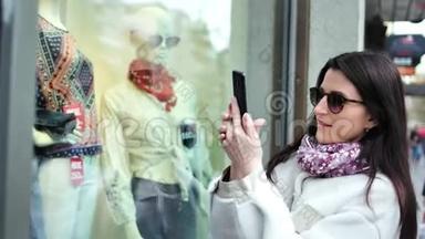 时尚的购物者女士用智能手机在商店时尚服装橱窗中拍照