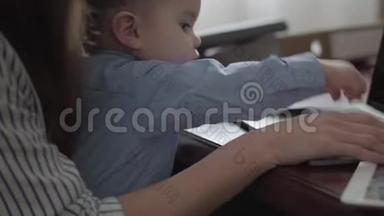 不认识的年轻母亲正拿着笔记本电脑和文件夹在桌子上工作。 小男孩坐着