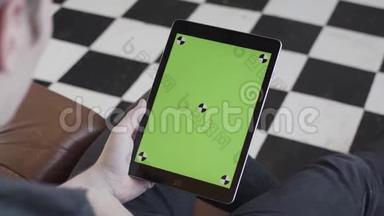 穿着深色牛仔裤的男人坐在沙发上，手里拿着平板电脑，<strong>黑白格子</strong>地板上有绿色屏幕