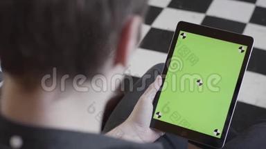 穿着深色牛仔裤的男人坐在沙发上，手里拿着平板电脑，<strong>黑白格子</strong>地板上有绿色屏幕