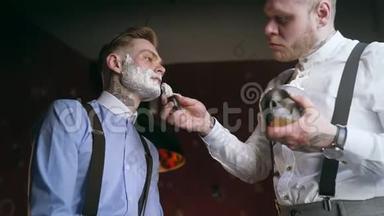 纹身理发师在他纹身的客户脸上用剃毛刷涂泡沫，30`黑手党风格的理发店，刮胡子