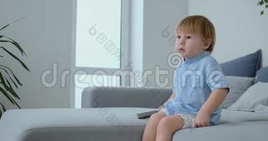 一个2岁的男孩坐在沙发上看电视，手里<strong>拿</strong>着<strong>遥控</strong>器。