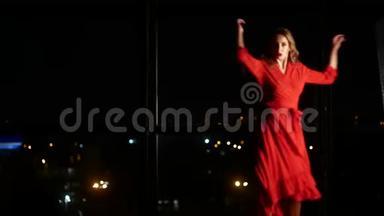 一个穿着红色优雅连衣裙的女孩正在一扇大窗户和一个夜晚的城市的背景下跳舞。 独自在家和